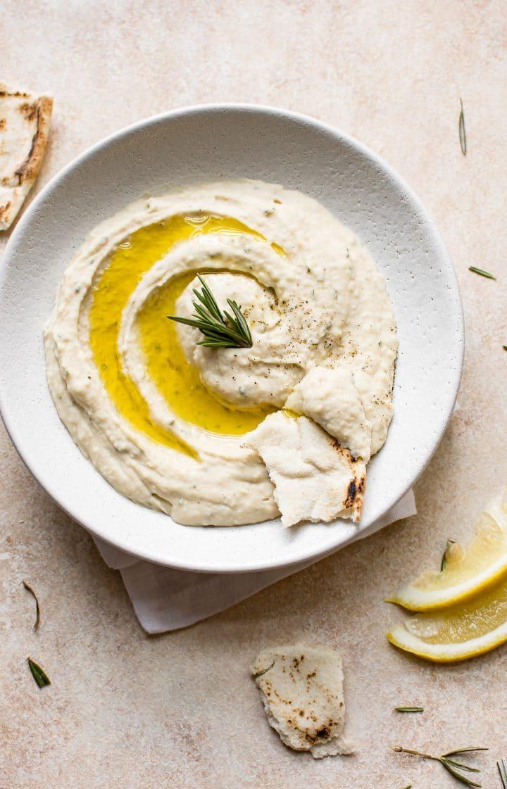 Veganer weißer Bohnen-Dip mit Rosmarin, Zitronensaft, Knoblauch und hochwertigem Olivenöl in einer weißen Schale, dazu frisches Pitabrot.
