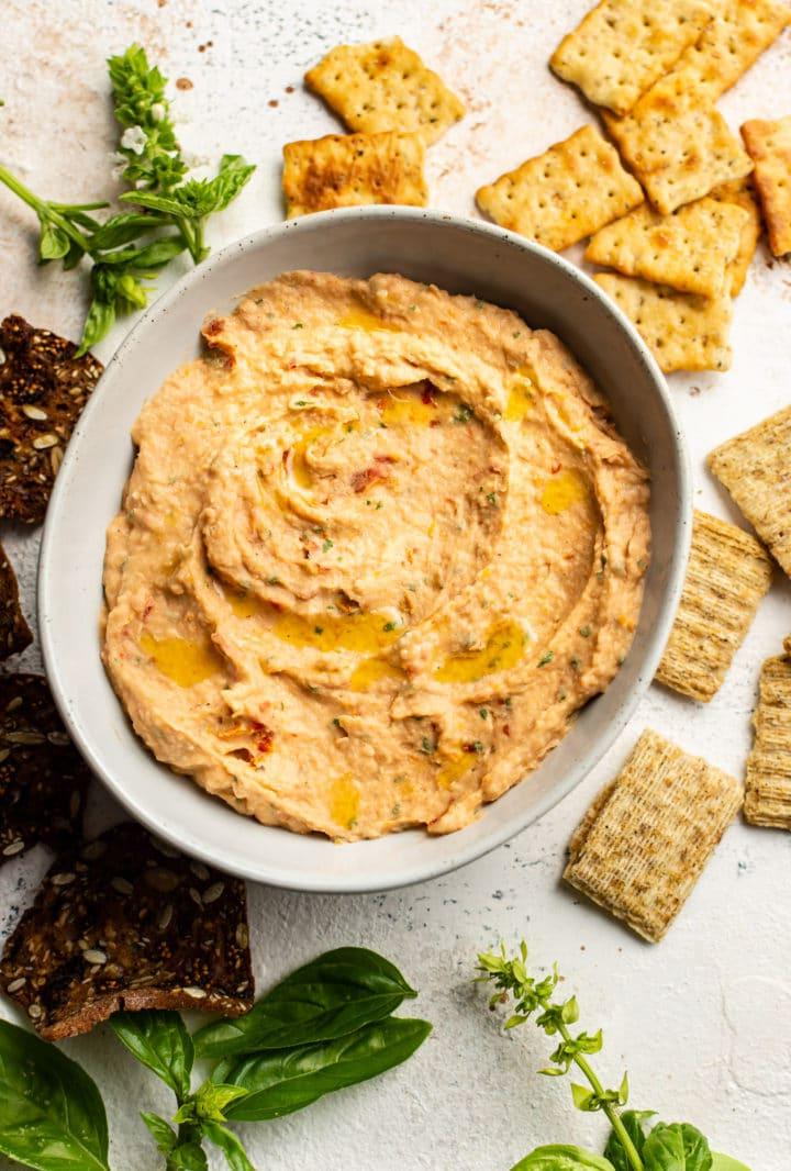 **Weißer Bohnen-Dip mit sonnengetrockneten Tomaten und Basilikum** 2 Weiße Bohnen Dip mit getrockneten Tomaten und Basilikum in einer Servierschale, umgeben von Crackern und frischem Basilikum