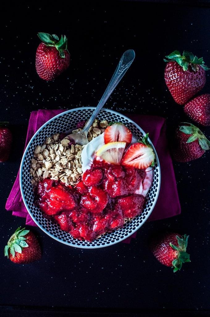 Schale mit Granola, Griechischem Joghurt und Erdbeer-Zitronen-Vanille-Soße mit einem Löffel und ganzen Erdbeeren daneben.