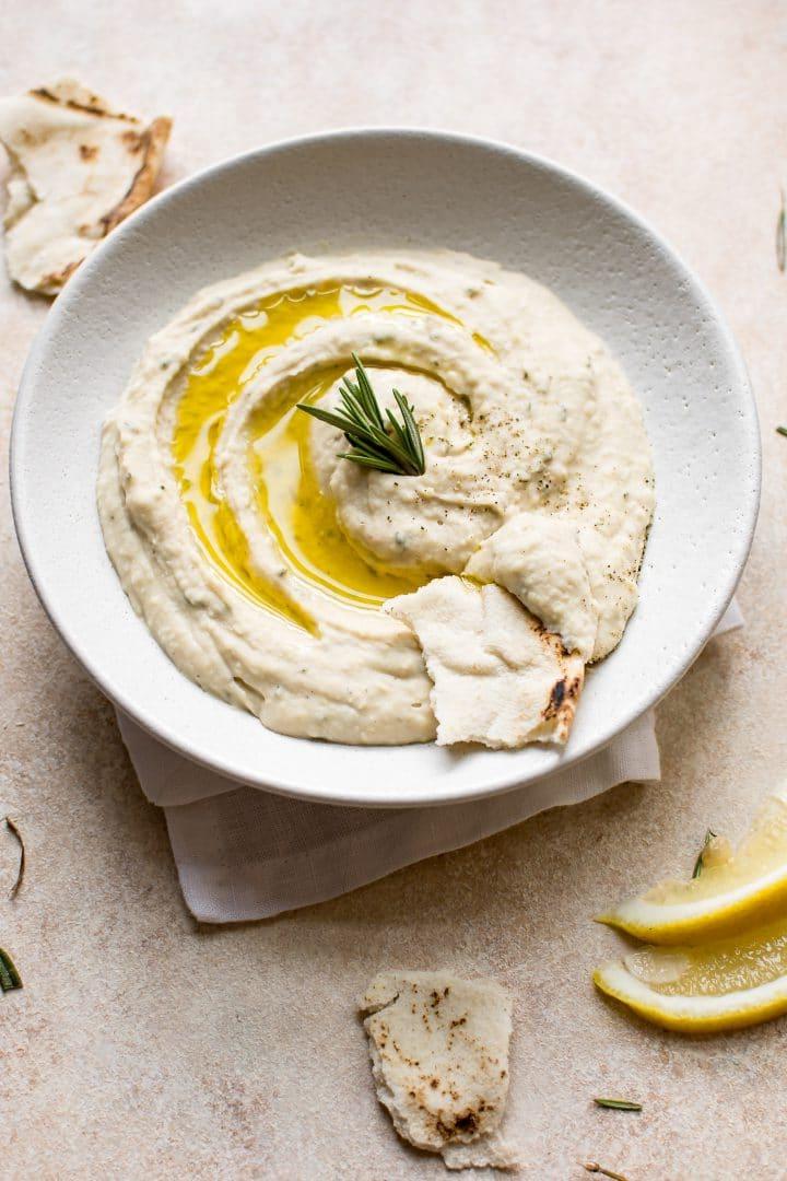 Veganer weißer Bohnen-Rosmarin-Dip in einer Schale, kunstvoll garniert mit einem Rosmarinzweig und serviert mit frischem Pitabrotstücken.