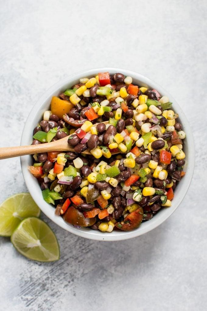 Weiße Schüssel mit schwarzem Bohnen- und Mais-Salat mit hölzernem Servierlöffel und Limettenspalten