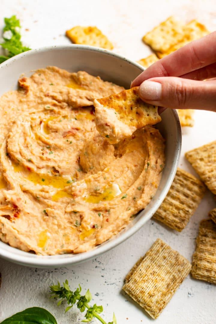 **Weißer Bohnen-Dip mit sonnengetrockneten Tomaten und Basilikum** 5 Nahaufnahme eines Crackers, der in den Cannellini-Bohnen-Dip getaucht wird