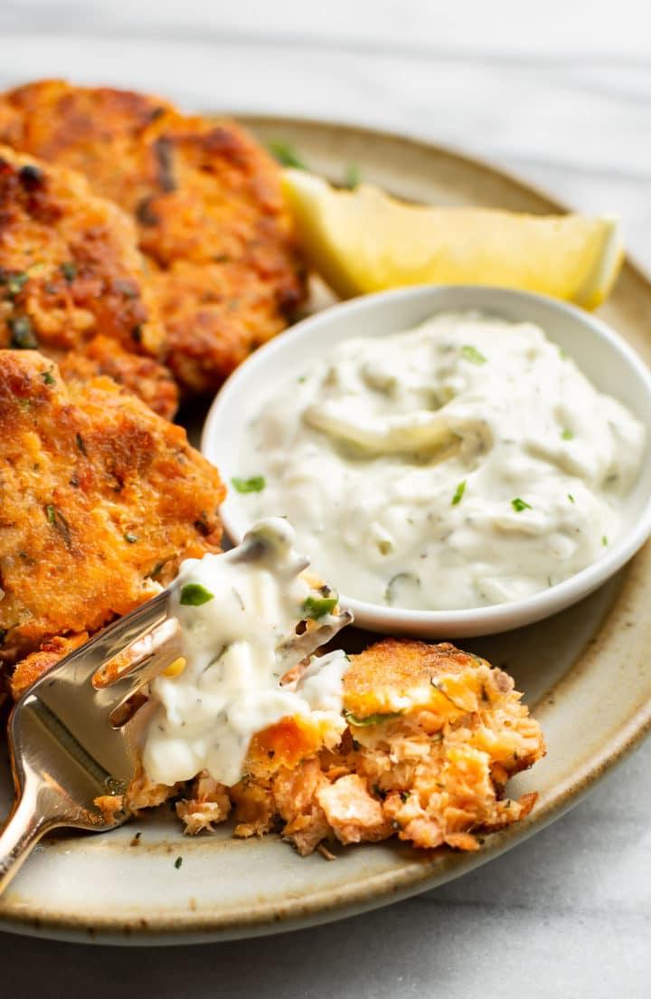 Nahaufnahme eines Tellers mit Lachs-Frikadellen, Dip und einer Zitronenspalte neben einer Gabel voll Lachs-Patty