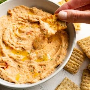 **Weißer Bohnen-Dip mit sonnengetrockneten Tomaten und Basilikum** 6 Nahaufnahme eines Crackers, der in den Cannellini-Bohnen-Dip getaucht wird