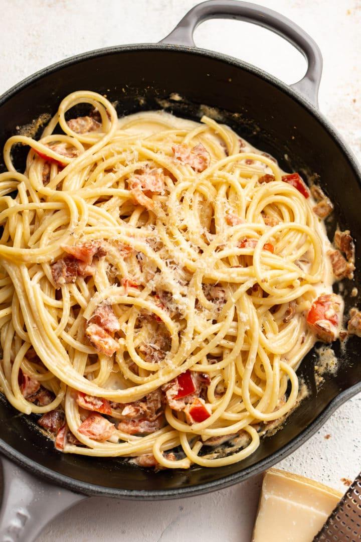 Einfache Cremige Speck-Tomaten-Nudeln 6 Speck- und Tomaten-Pasta in einer Pfanne mit einem Stück Parmesan und einer Käsereibe daneben