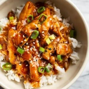 Honig-Knoblauch-Hähnchenschenkel aus dem Slow Cooker in einer Schüssel, über Reis serviert und mit Frühlingszwiebeln und Sesamkörnern garniert