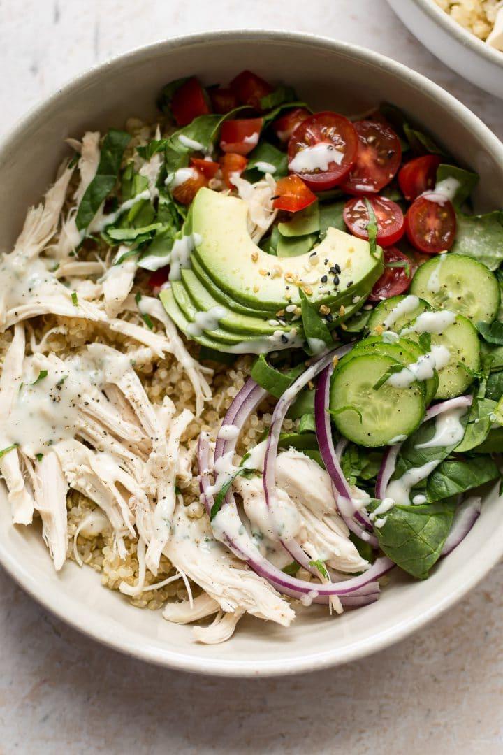 Weiße Schüssel mit Ranch Hähnchen-Quinoa-Salat, garniert mit Avocado, roten Zwiebeln, Tomaten, Gurken und einem hausgemachten Ranch Dressing, das den Geschmack hervorhebt.