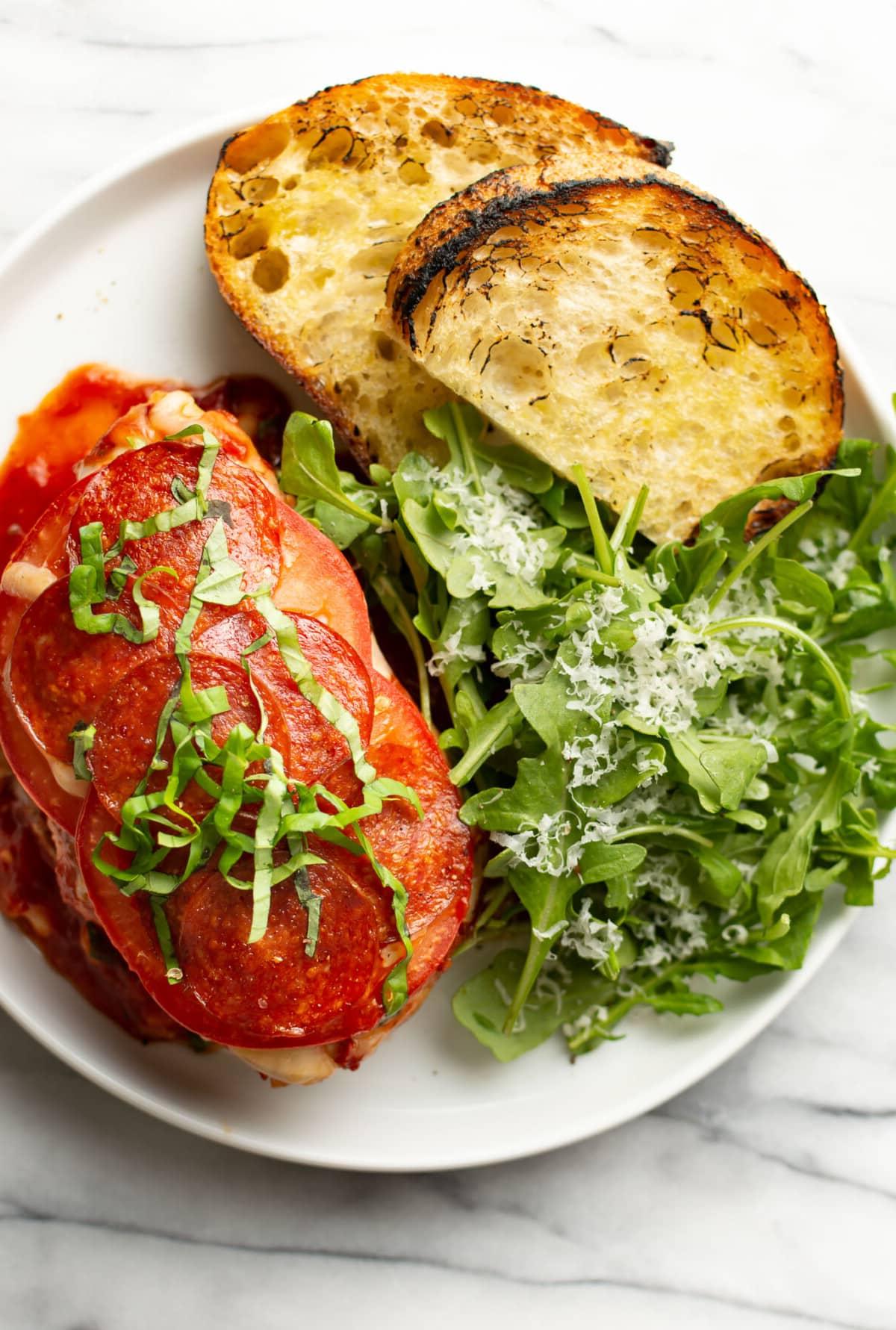 Pizza Hähnchen 8 Ein angerichteter Teller mit Pizza-Hähnchen, geröstetem Brot und einem Rucola-Salat als Beilage