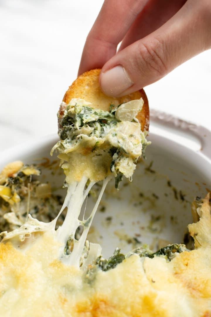 Heißer Spinat-Artischocken-Dip 8 Nahaufnahme einer Hand, die Brot in Spinat-Artischocken-Dip taucht
