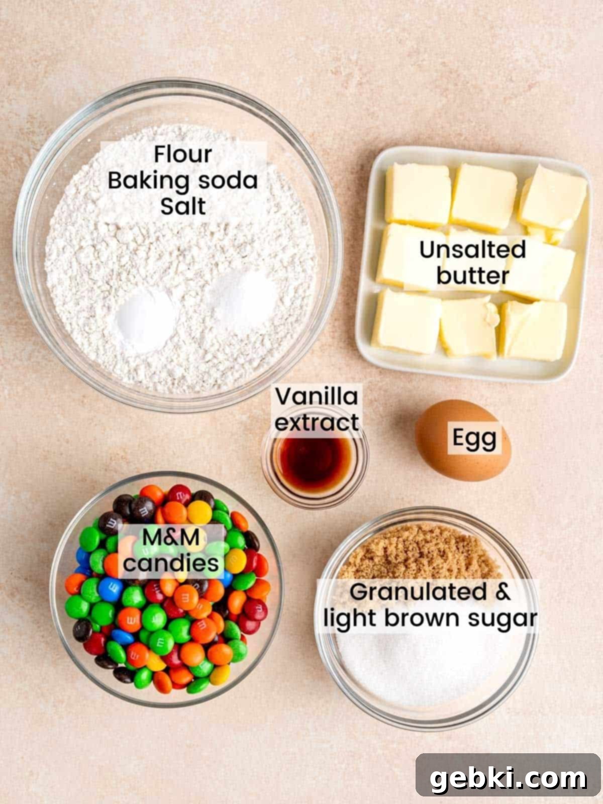 A selection of ingredients laid out on a white surface, including butter, sugars, eggs, flour, and M&M candies, ready for making soft M&M sugar cookies.
