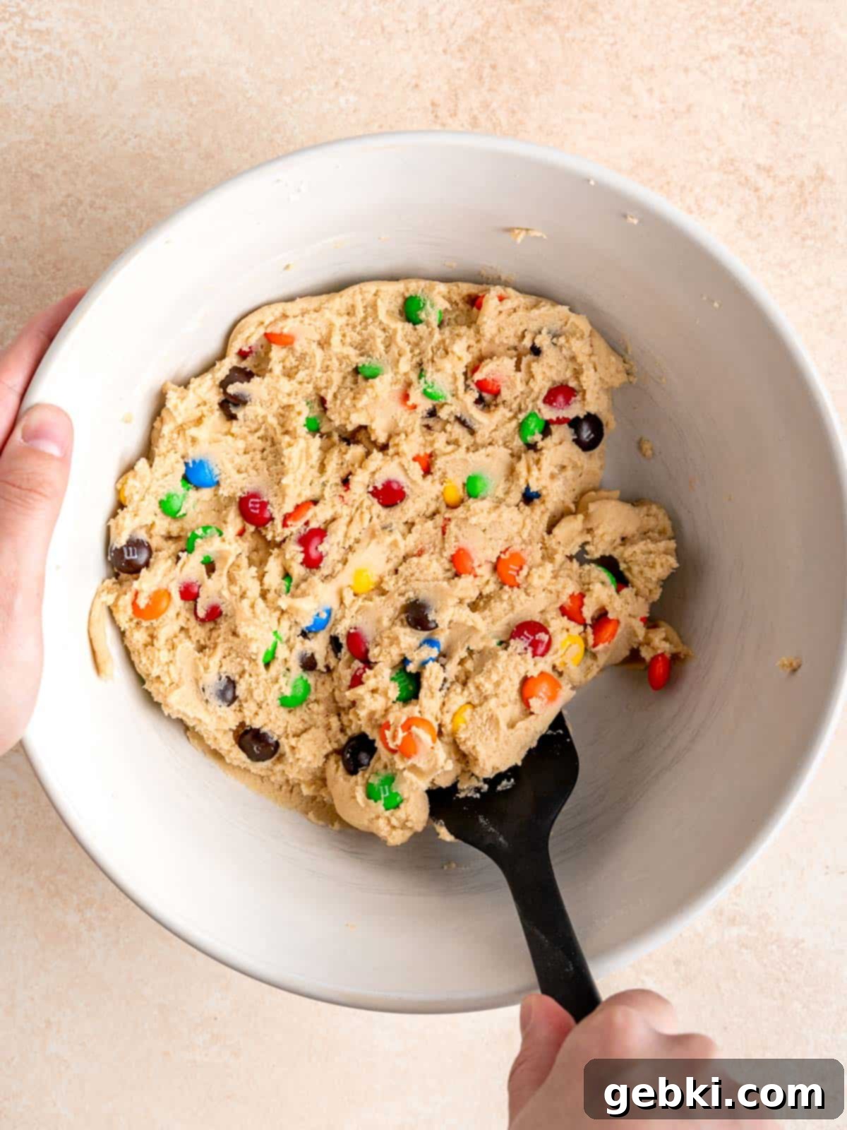 M&Ms being gently mixed into sugar cookie dough with a rubber spatula to prevent breaking.