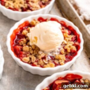 A golden-brown strawberry rhubarb crisp in a ramekin, with a scoop of vanilla ice cream melting on top, seen from a slightly elevated angle.
