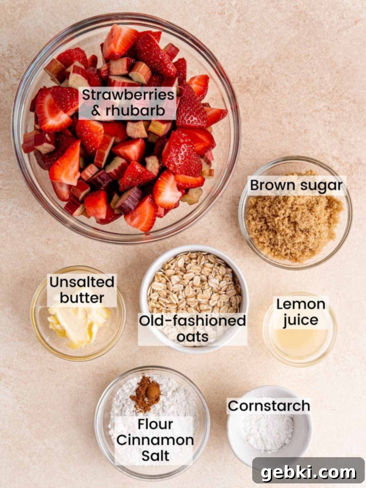 A collection of fresh ingredients laid out, including chopped strawberries and rhubarb, light brown sugar, cornstarch, lemon juice, butter, oats, and flour, ready for making strawberry rhubarb crisp.