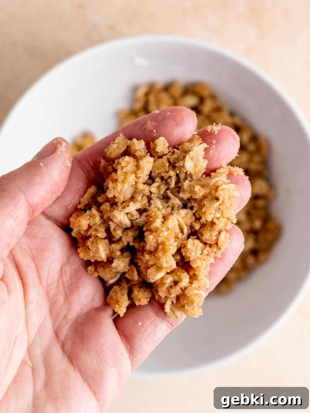 A hand mixing a crumbly blend of brown sugar, flour, melted butter, oats, cinnamon, and salt in a bowl, showing the perfect texture for the crisp topping.