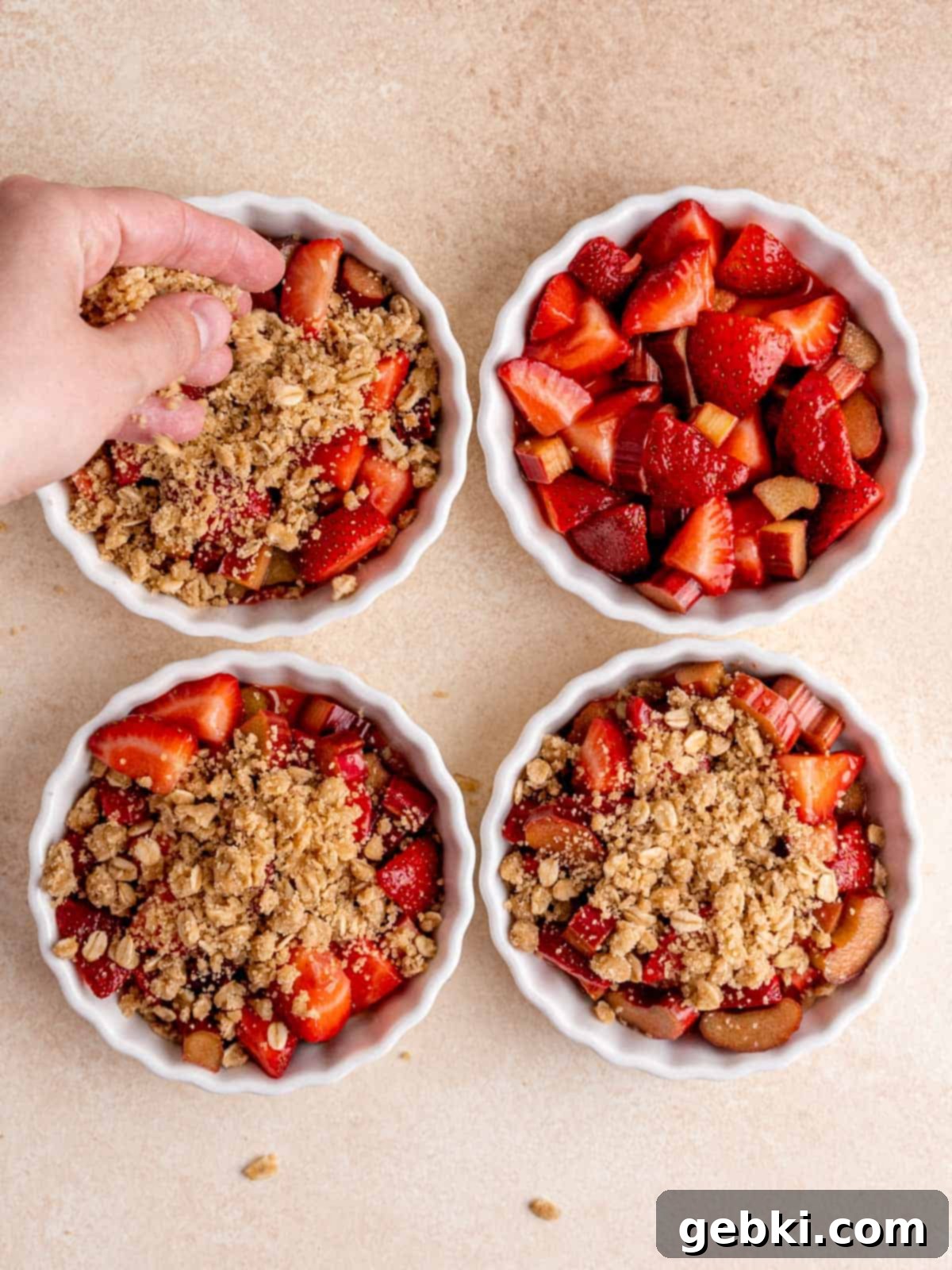 A hand sprinkling the buttery oat crisp topping evenly over the strawberry rhubarb fruit filling in individual ramekins, ready for baking.