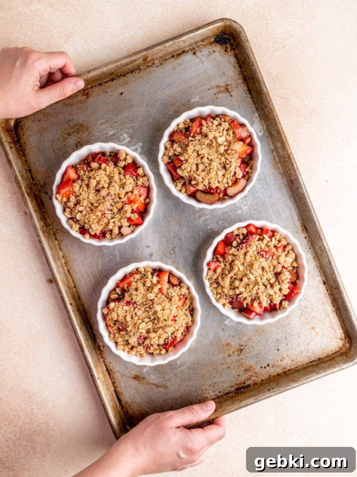 Four individual ramekins of unbaked strawberry rhubarb crisp with oat topping, placed on a baking sheet, ready to go into the oven.