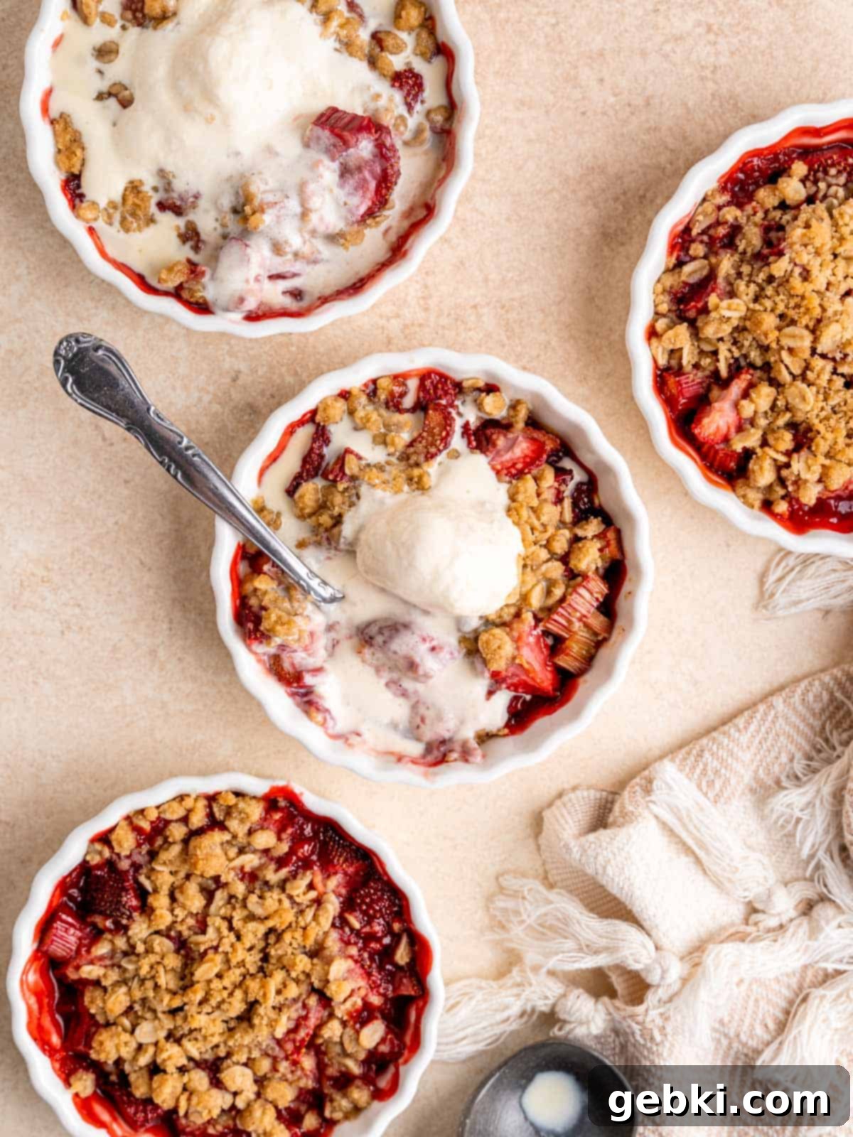 Close-up top view of two baked strawberry rhubarb crisps in ramekins, both topped with melting vanilla ice cream, showcasing the golden oat topping and bubbly fruit.