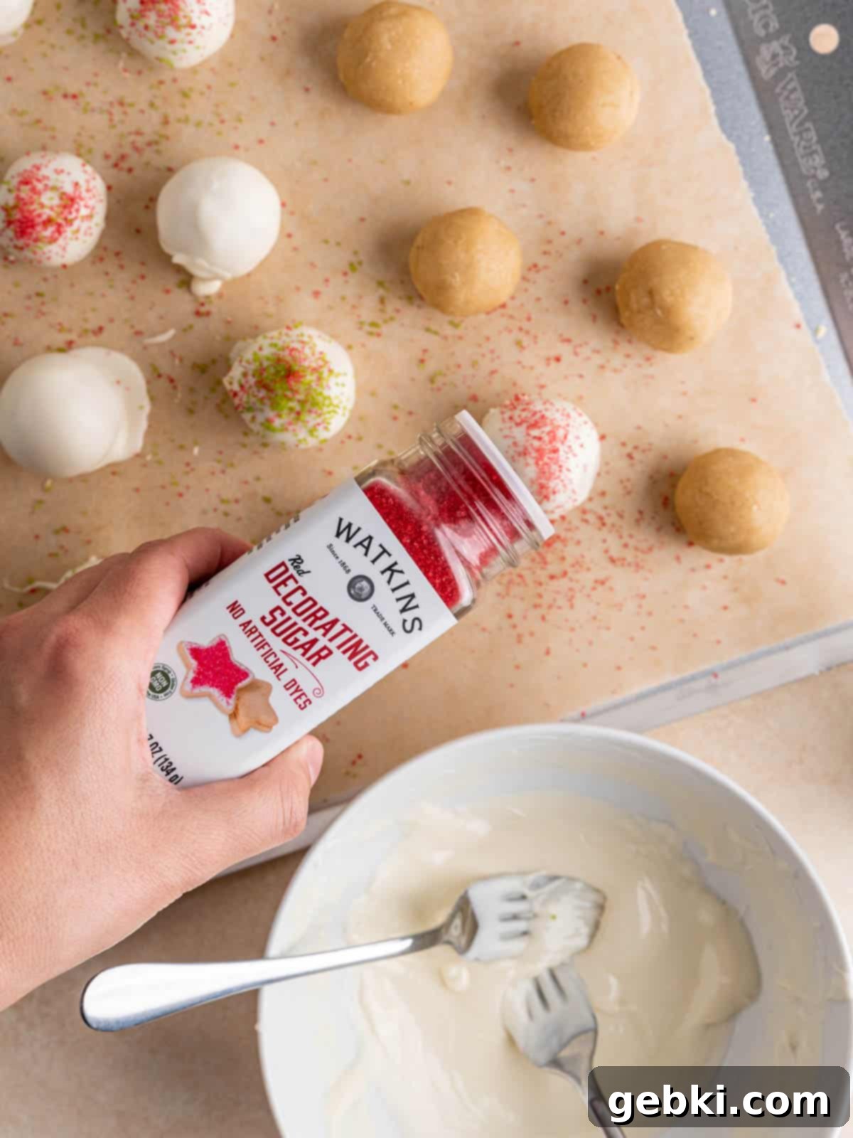 No-Bake Christmas Cookie Truffles 11 Watkins red decorating sugar being artfully sprinkled over a freshly white chocolate-covered sugar cookie truffle for a vibrant holiday finish.