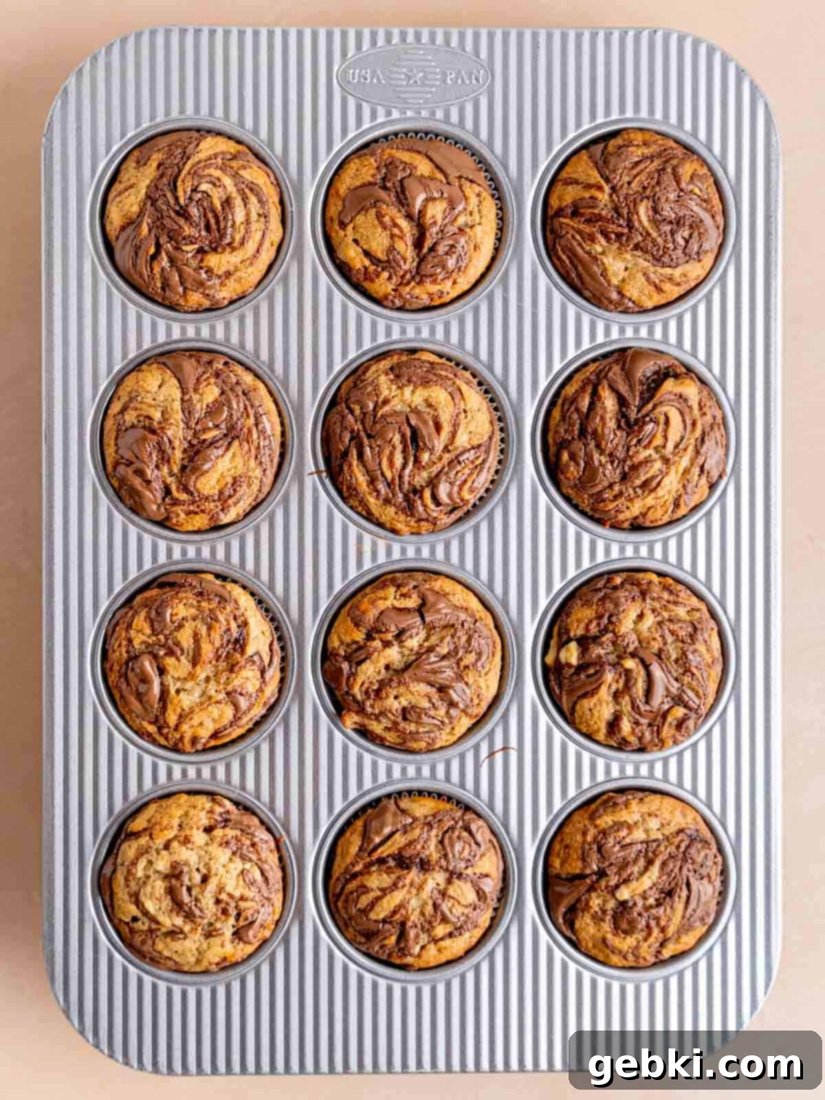 Marbleized Banana Nutella Muffins 9 Freshly baked banana Nutella muffins cooling in a muffin pan, their golden tops and visible, enticing swirls indicating their deliciousness and readiness to be enjoyed.