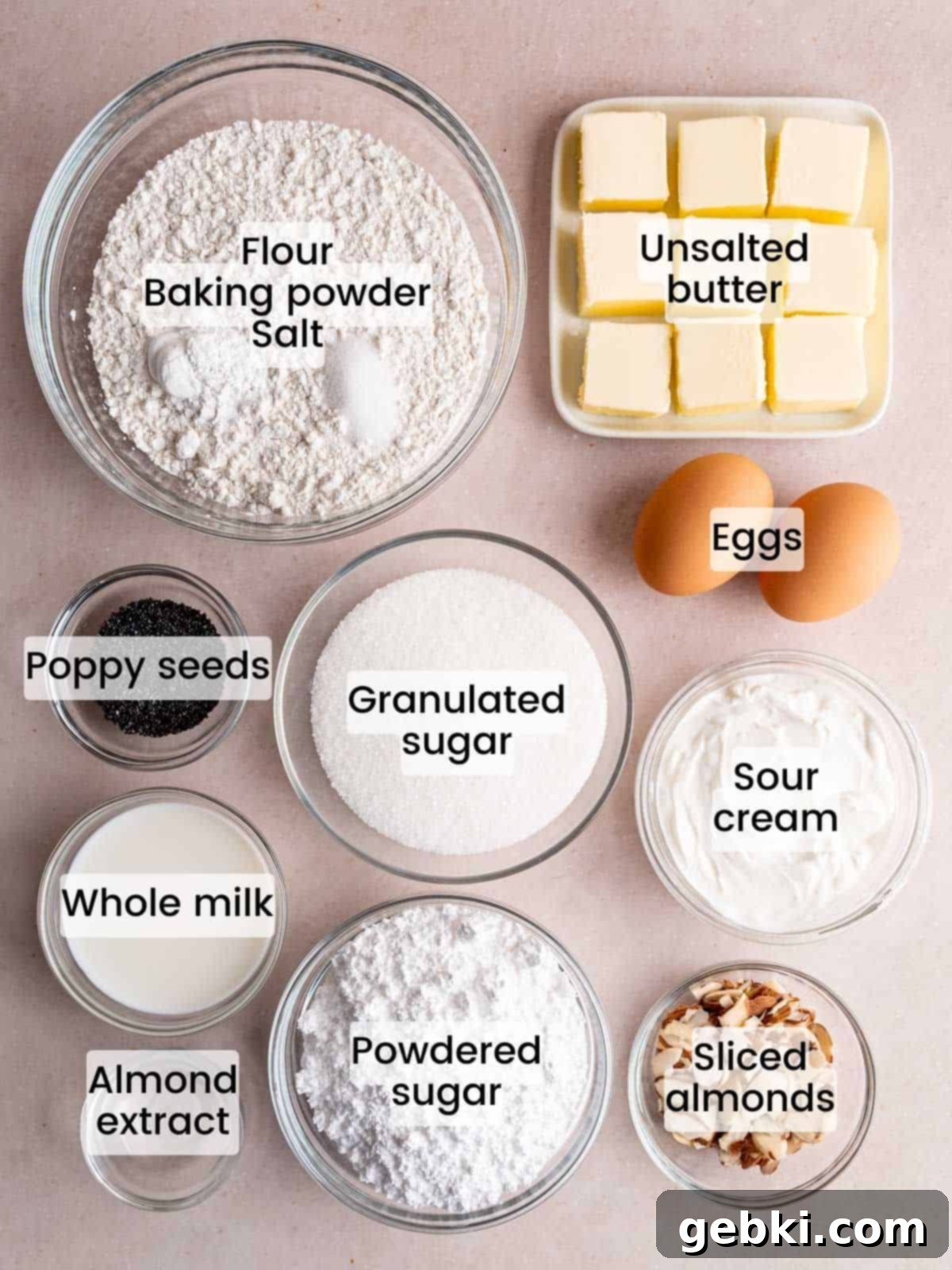A collection of fresh ingredients laid out, including flour, sugar, butter, eggs, sour cream, almond extract, milk, poppy seeds, and sliced almonds, ready to make a glazed almond poppy seed loaf cake.