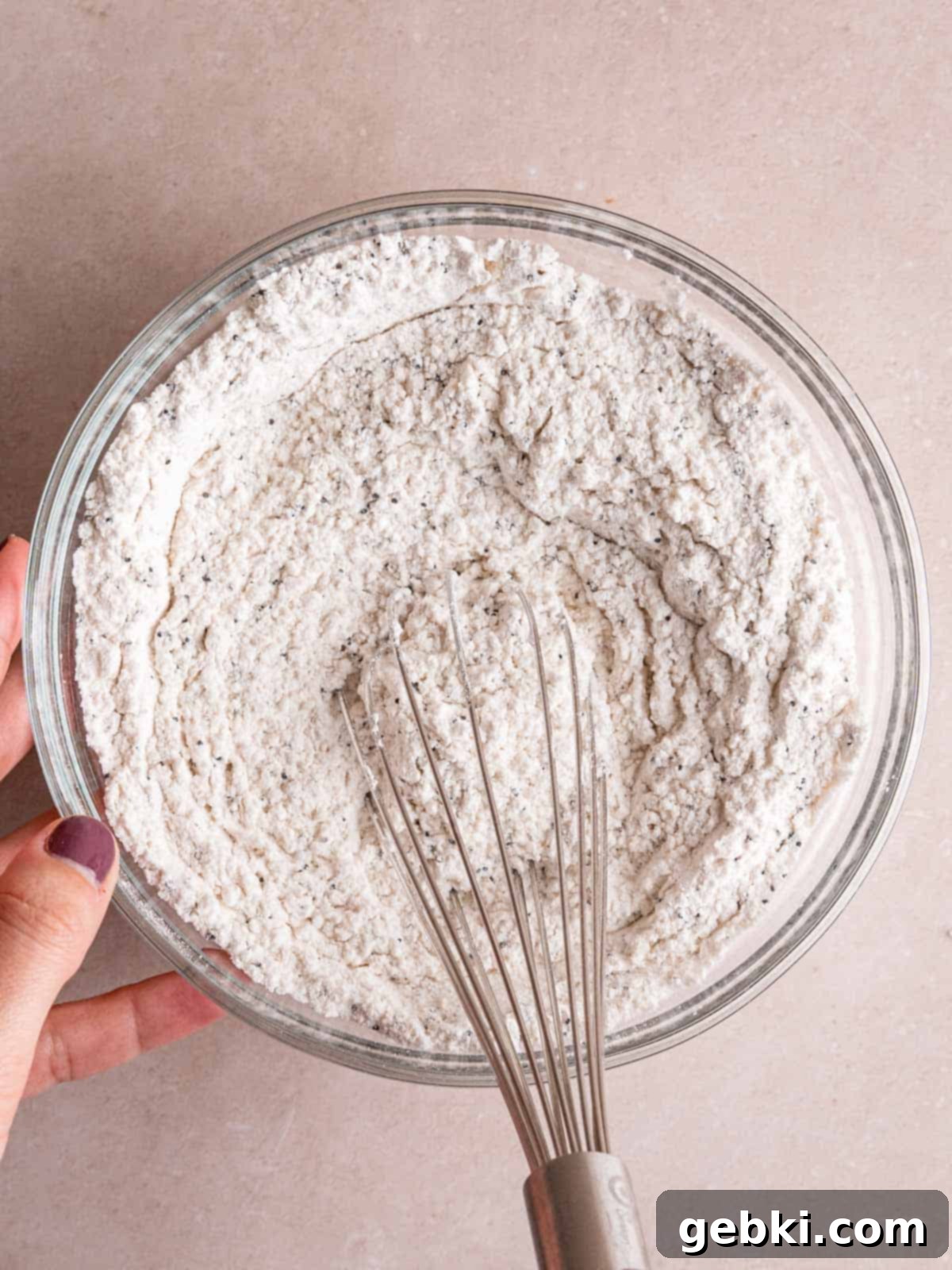 A medium bowl containing all-purpose flour, baking powder, salt, and poppy seeds, being whisked together until thoroughly combined.