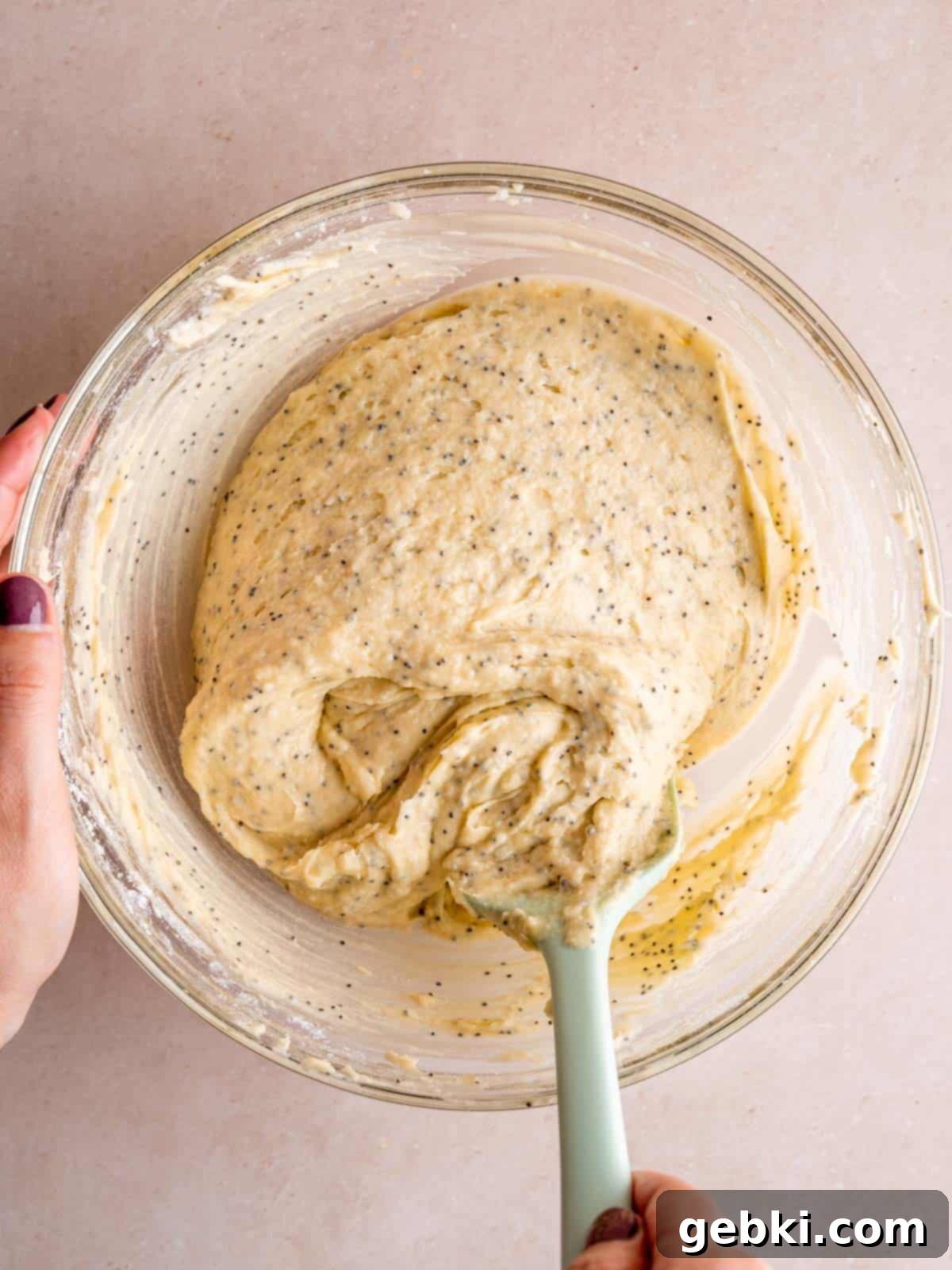 Smooth almond poppy seed cake batter in a mixing bowl, with a spatula demonstrating its perfect consistency after mixing wet and dry ingredients.