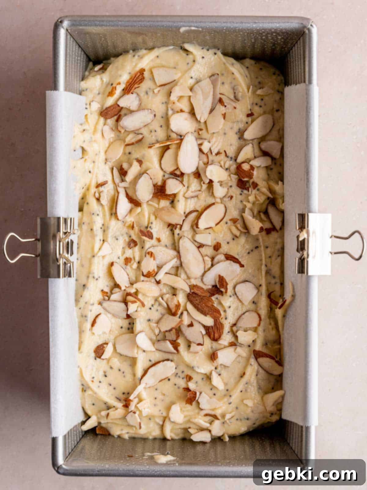Almond poppy seed cake batter evenly spread in a parchment-lined loaf pan, topped with a generous sprinkle of sliced almonds, ready for baking.