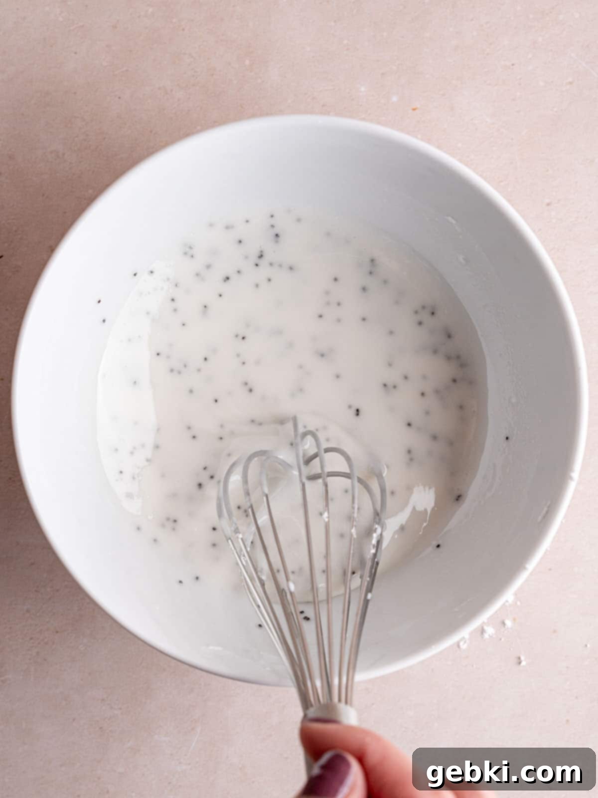 A small bowl holding a smooth, white almond-flavored glaze, speckled with poppy seeds, being whisked to perfection.