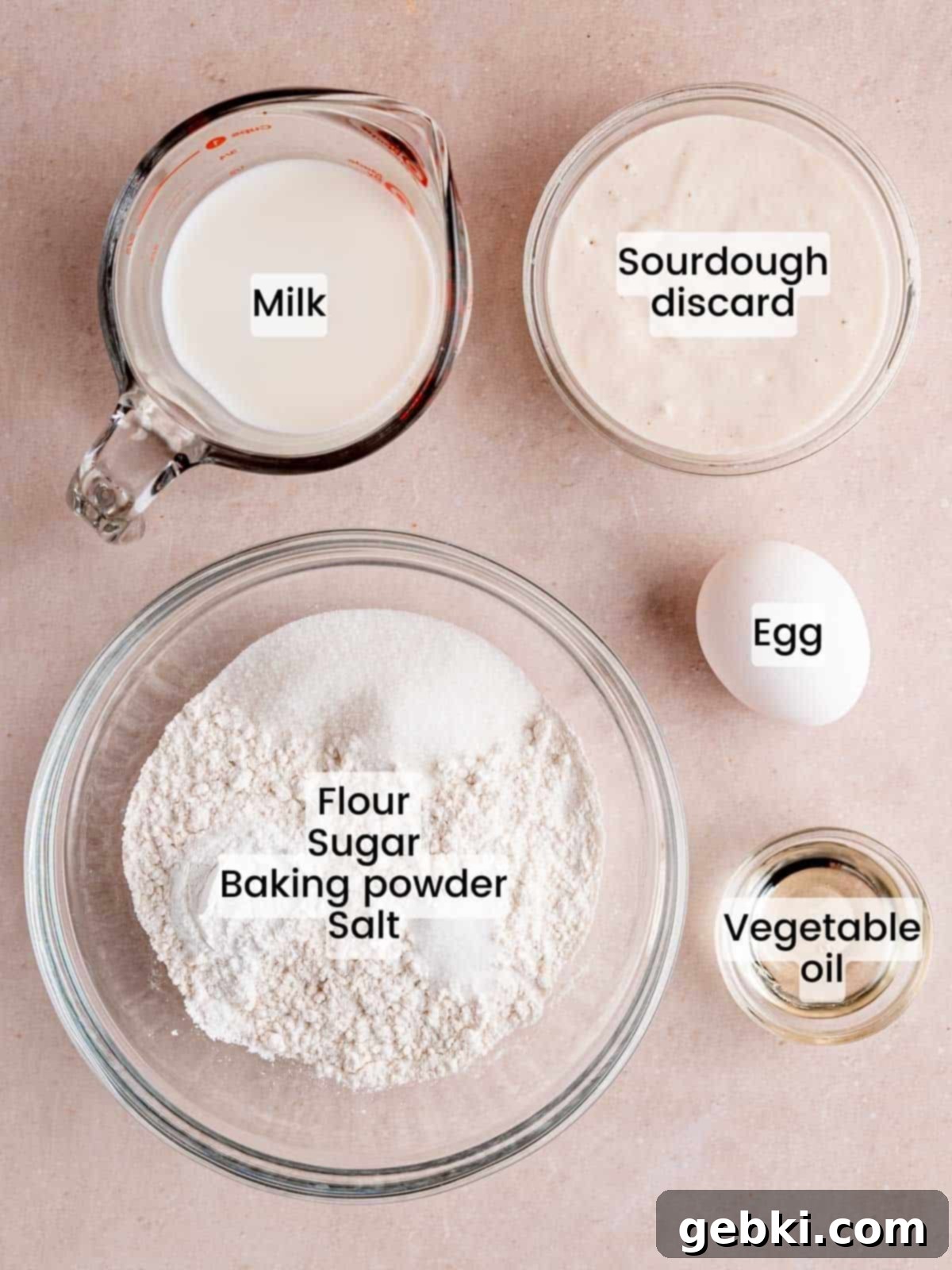 Turn Sourdough Discard Into Fluffy Pancakes 3 A flat lay of fresh ingredients laid out on a clean kitchen counter, including a bowl of all-purpose flour, granulated sugar, baking powder, salt, a whole egg, a bottle of vegetable oil, a jar of sourdough discard, and a carton of milk, all ready for pancake preparation.