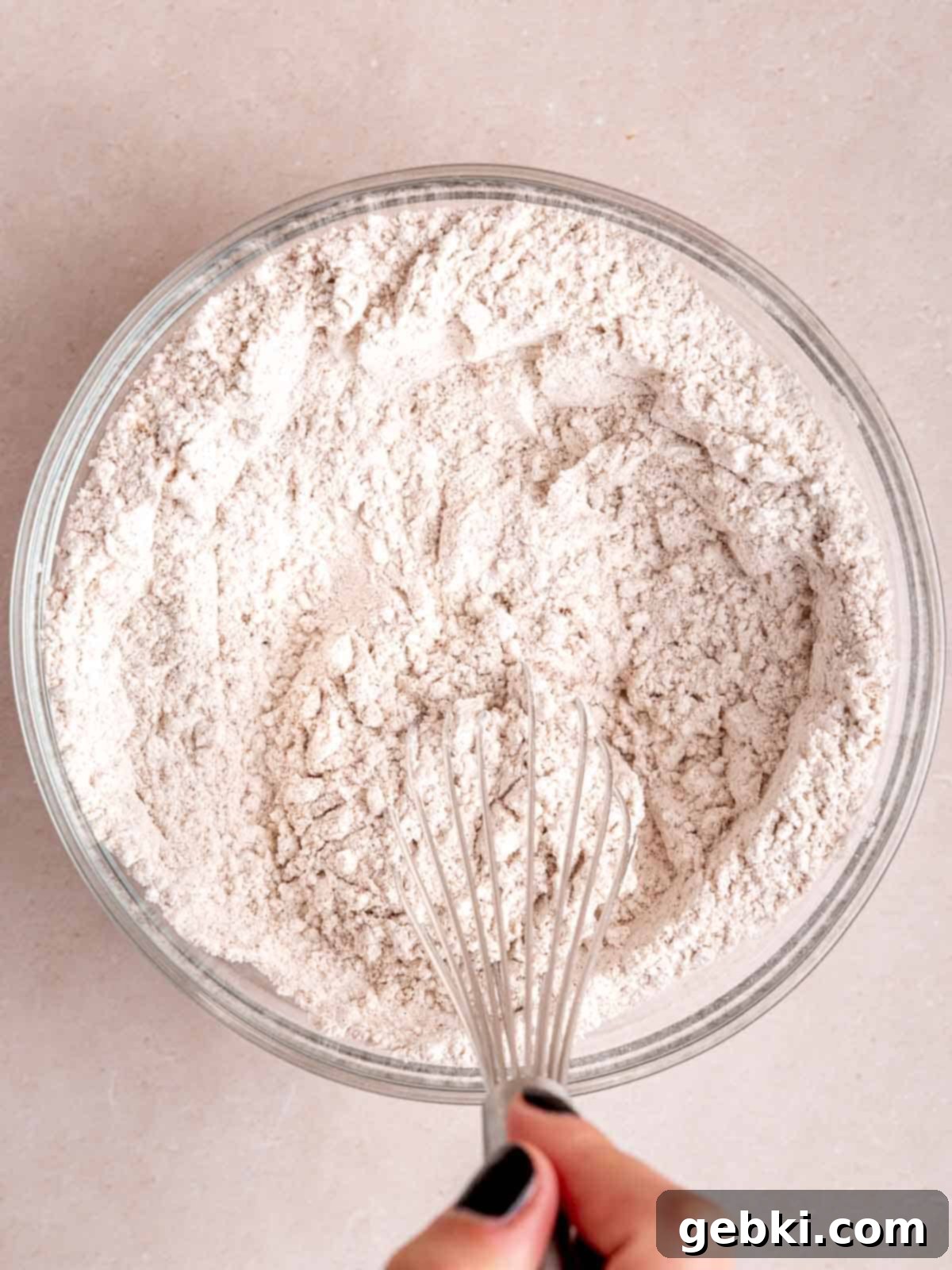 A hand whisking together dry ingredients: flour, baking powder, baking soda, cinnamon, and pumpkin pie spice in a large mixing bowl.