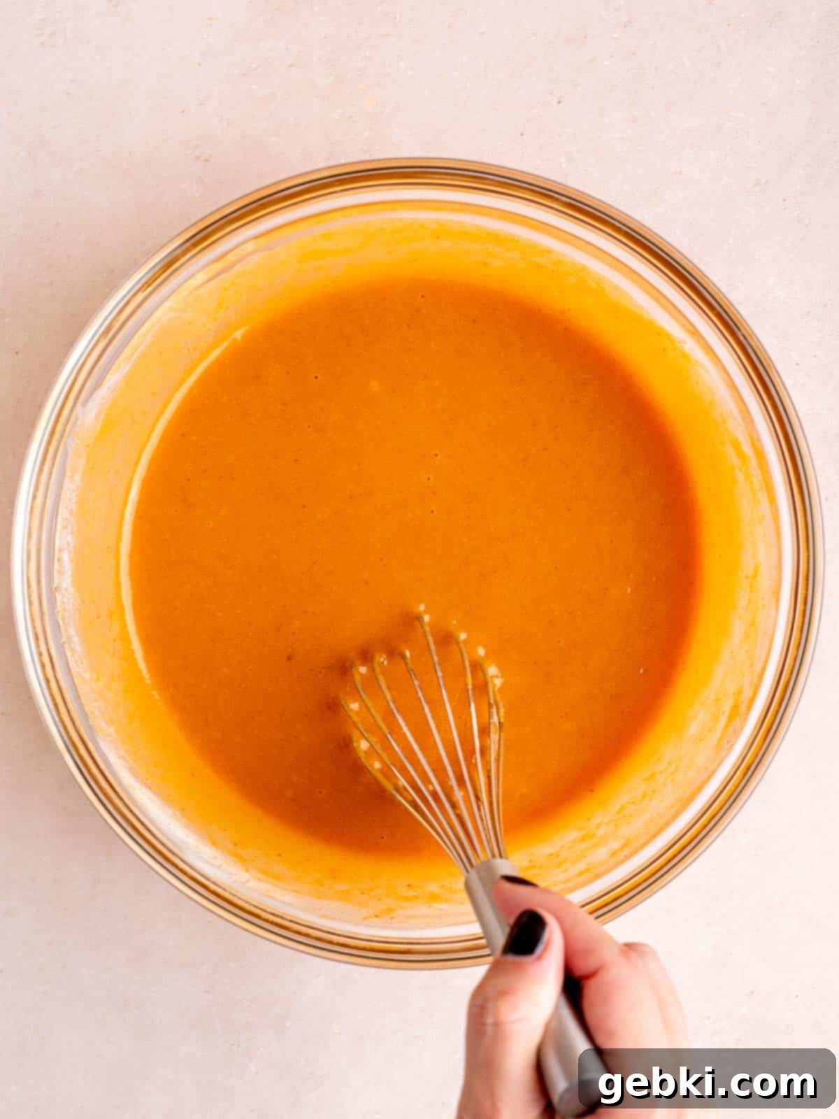 A whisk blending vegetable oil, granulated and brown sugar, eggs, vanilla extract, kefir, and pumpkin puree in a large bowl to form the wet ingredients for pumpkin muffins.