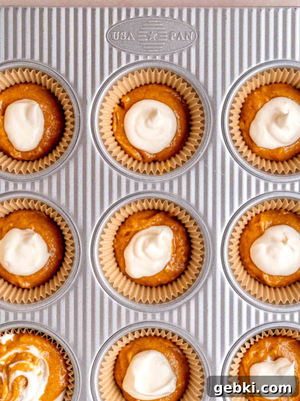 A muffin pan with liners partially filled with pumpkin batter and dollops of creamy cream cheese filling, showing the layering process.