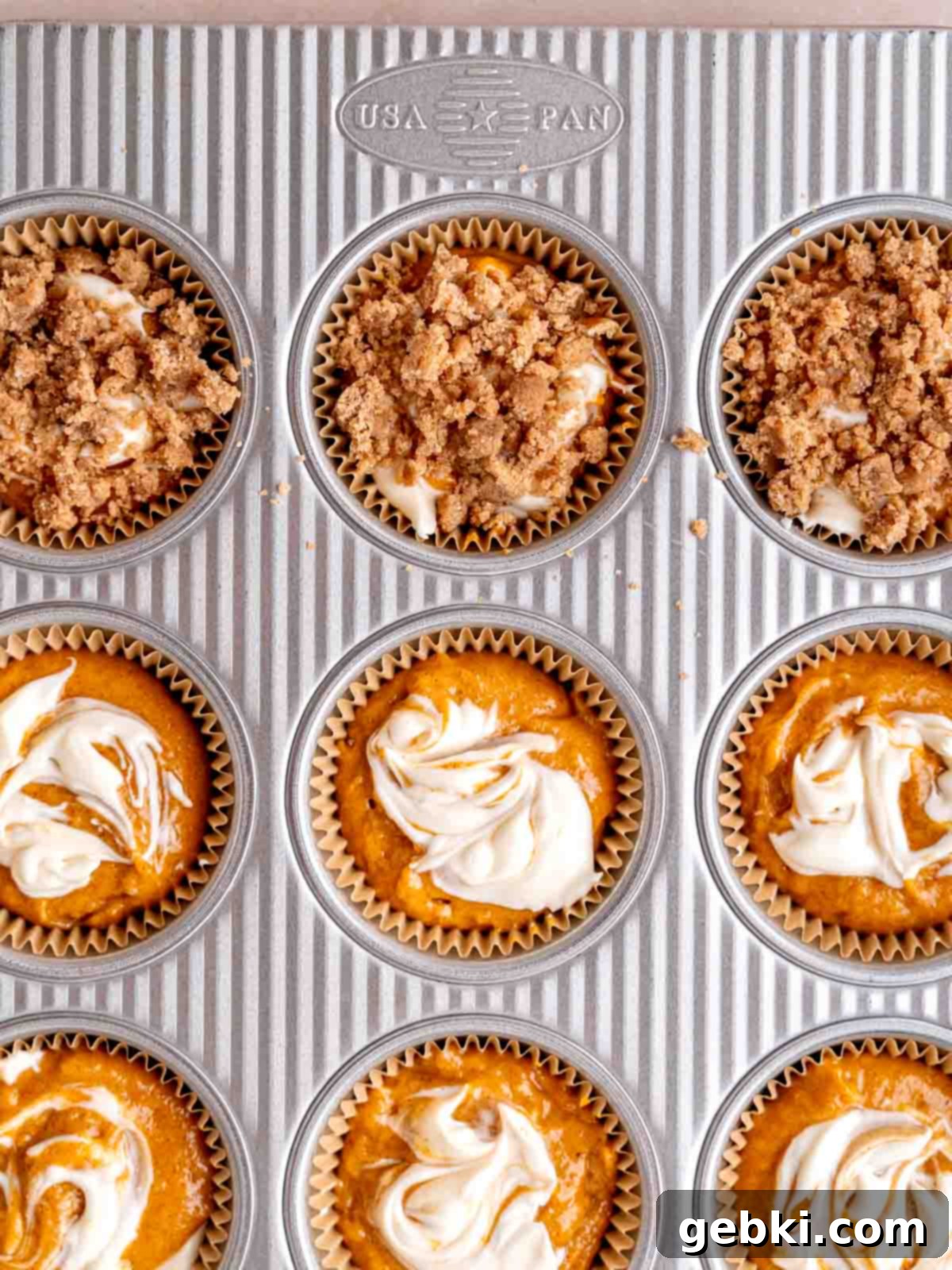 Muffin liners filled with layers of pumpkin batter, cream cheese filling, and a final swirl of cream cheese topped with cinnamon streusel, ready for baking.