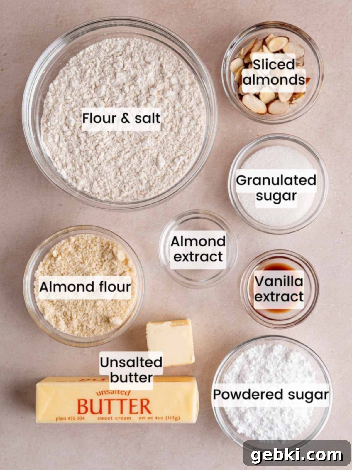 A flat lay photograph showcasing all the ingredients required to make delicious almond croissant shortbread cookies, including butter, sugars, extracts, flours, and sliced almonds.