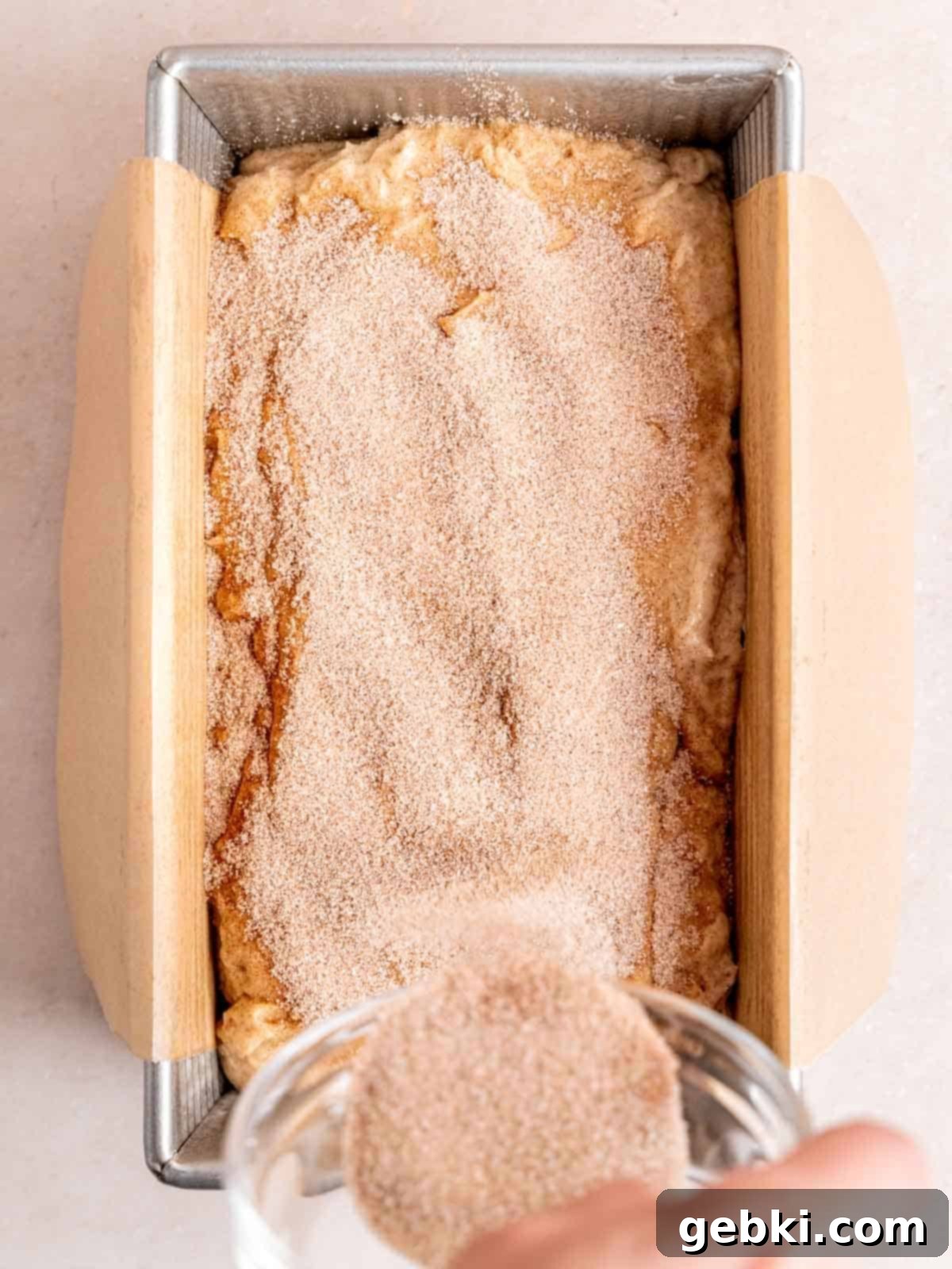 Sprinkling a cinnamon sugar topping evenly over cinnamon cake batter in a parchment-lined loaf pan before baking.