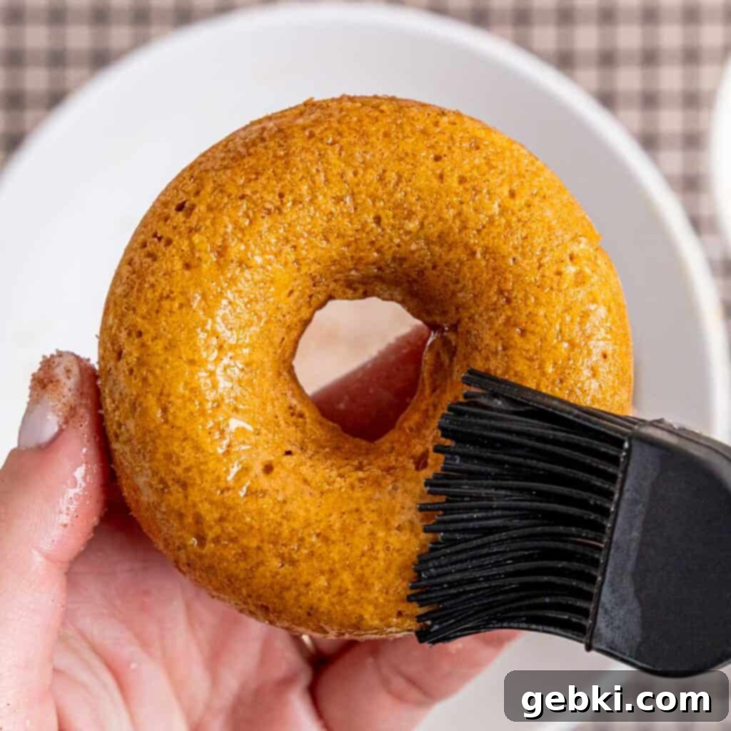 Brushing melted butter onto a baked pumpkin donut to prepare it for cinnamon sugar coating.