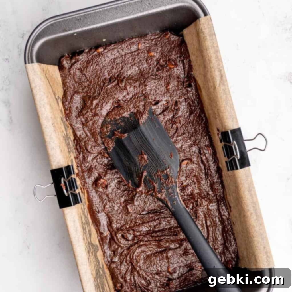 Spreading chocolate chip brownie batter into a loaf pan using a spatula.