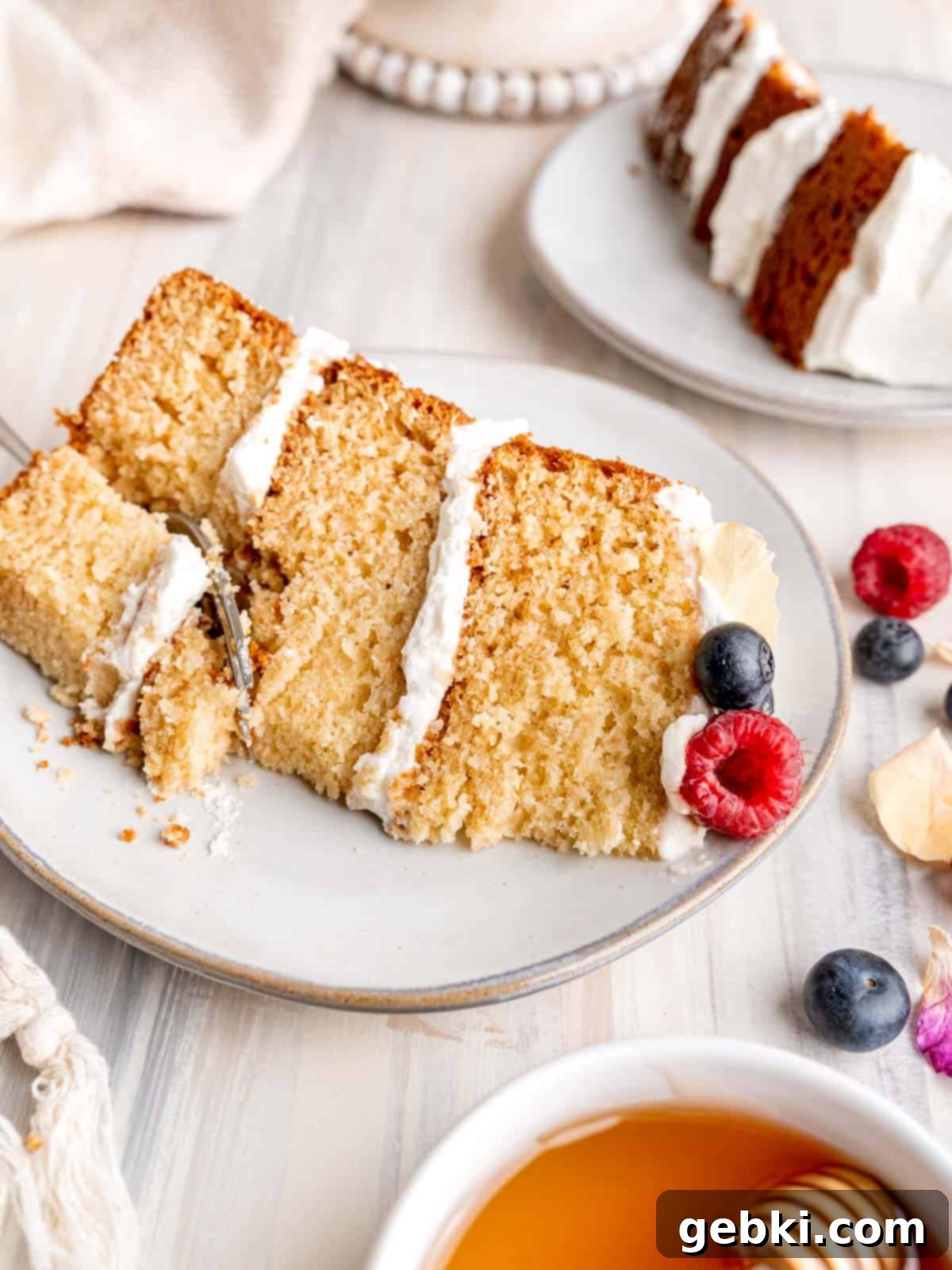 Effortless Honey Vanilla Delight 10 A fluffy slice of honey vanilla cake with whipped cream, topped with fresh berries and a drizzle of honey, on a white plate.