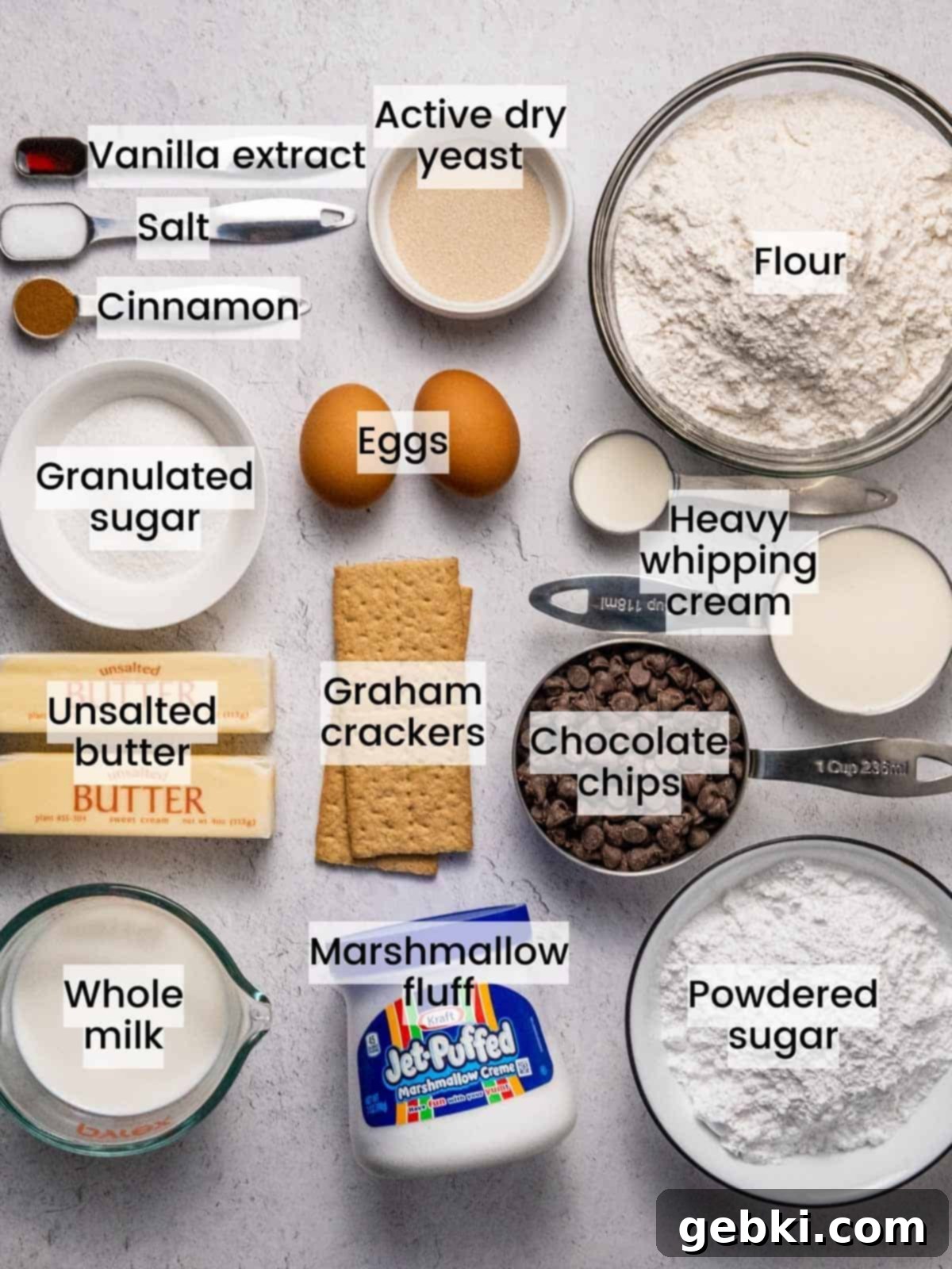 Gooey S'mores Stuffed Donuts 2 Various ingredients for S'mores donuts laid out on a kitchen counter, including milk, yeast, butter, sugar, vanilla, salt, cinnamon, eggs, flour, vegetable oil, graham crackers, marshmallow fluff, powdered sugar, heavy whipping cream, and chocolate chips.