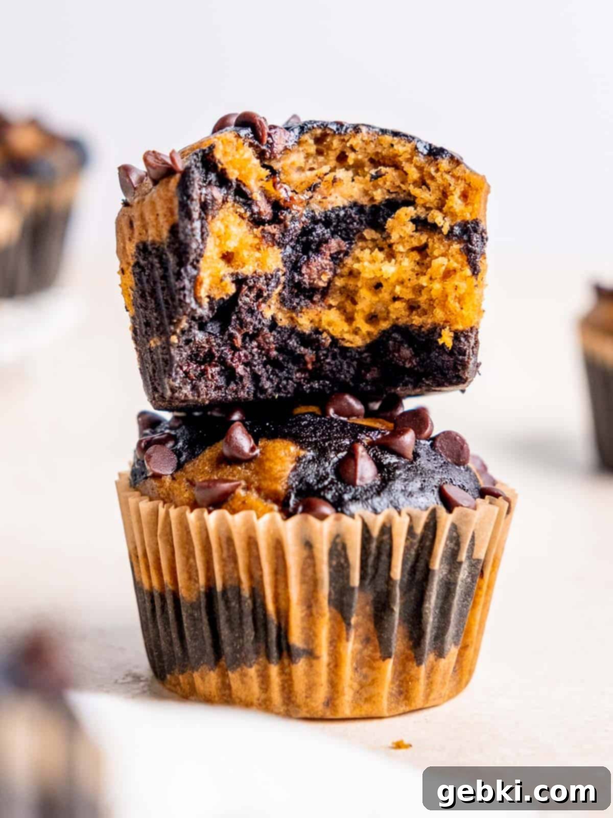 One unwrapped chocolate pumpkin muffin with a bit taken out sitting on top of another muffin in its liner, showcasing the marbled effect and rich colors.