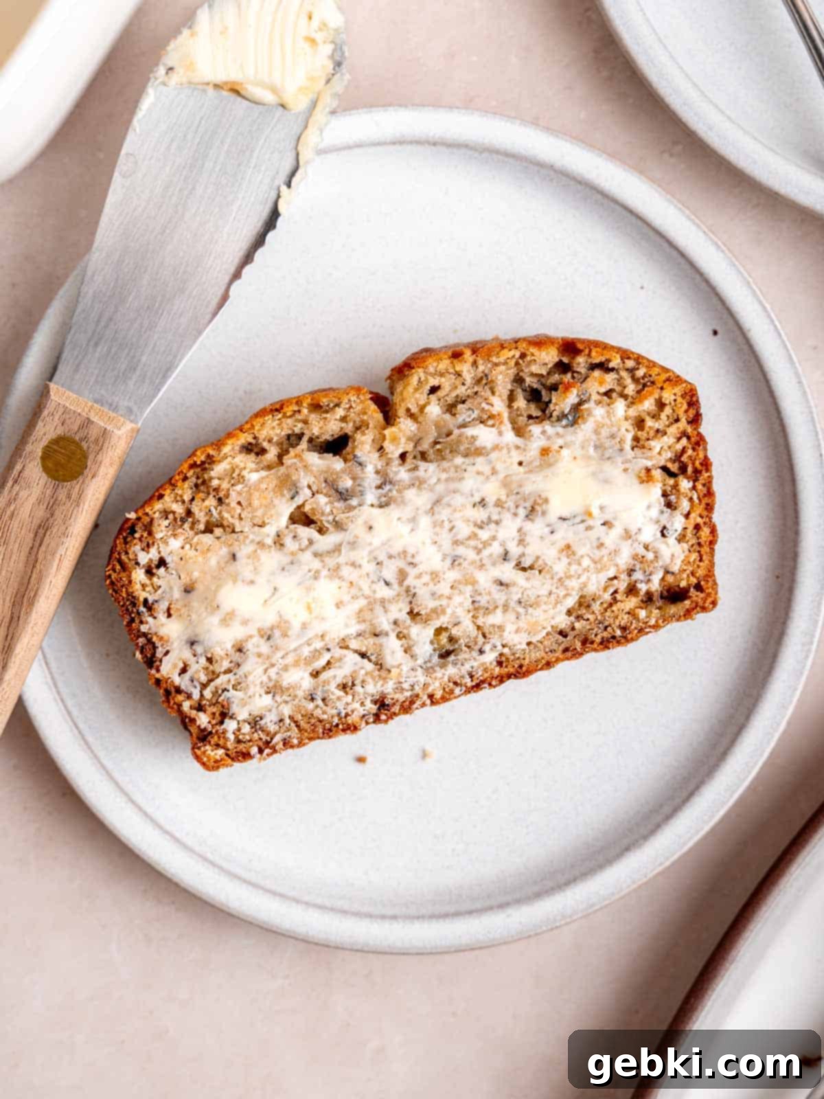 Effortless One-Bowl Banana Bread 8 A slice of banana bread covered in butter on a plate with a butter knife.