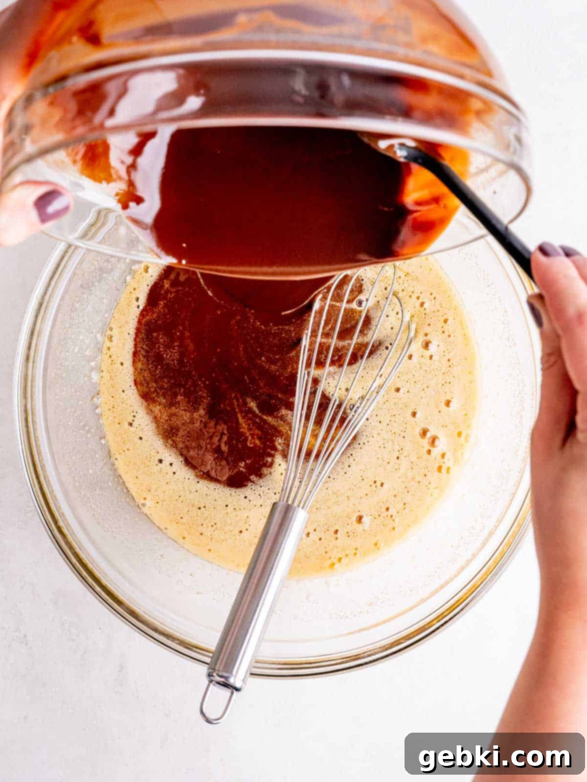 Pouring a slightly cooled melted chocolate mixture into a bowl of whisked eggs and sugars, being careful to mix gradually.