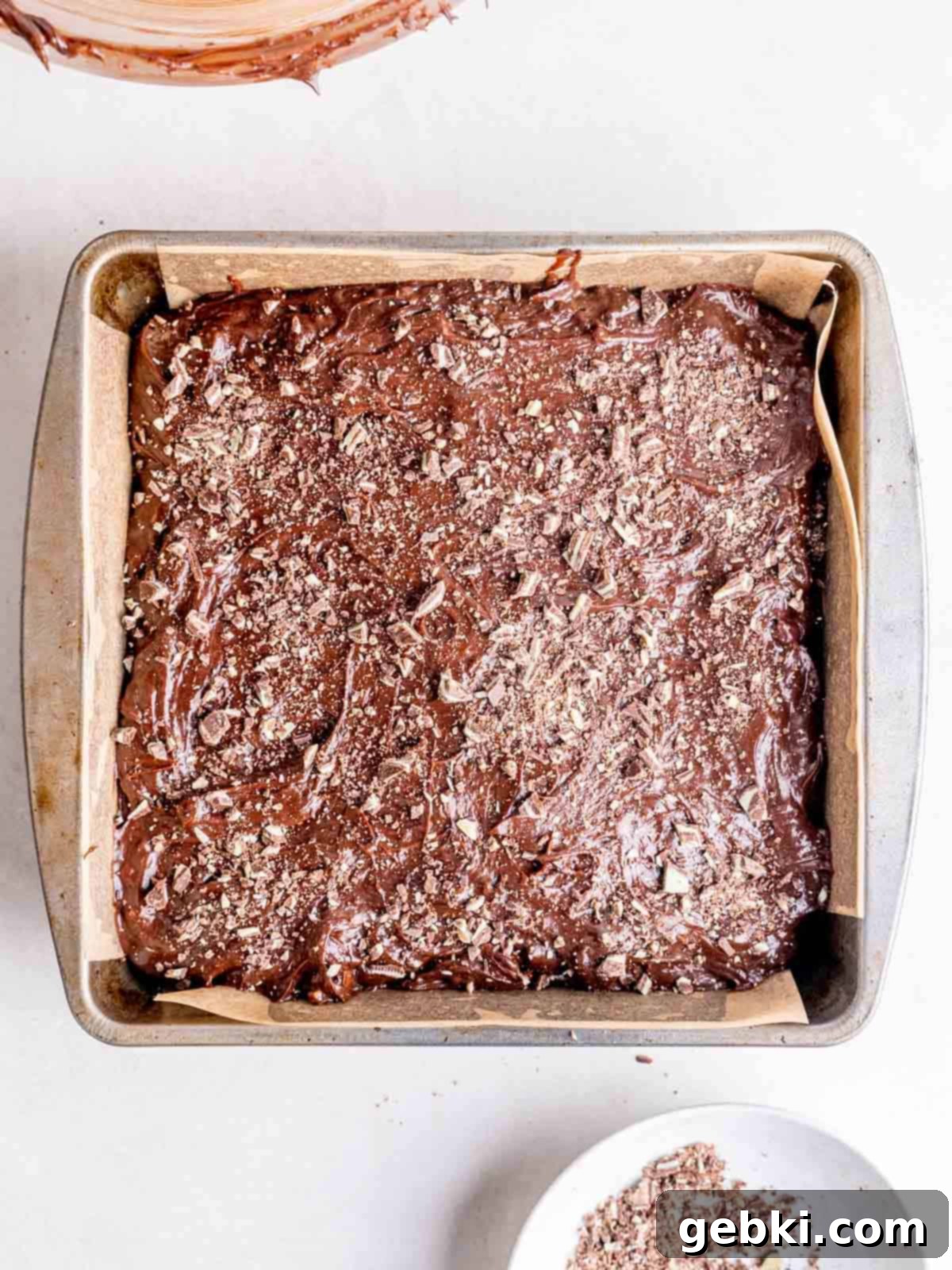 Mint chocolate brownie batter spread evenly in a parchment-lined pan, topped with additional finely chopped Andes mint pieces, ready for baking.