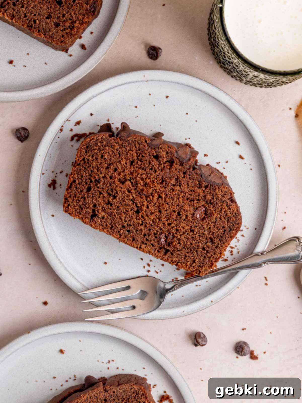 Decadent Double Chocolate Loaf 2 A beautifully presented slice of chocolate loaf cake on a pristine white plate, accompanied by a fork, ready to be enjoyed. The cake showcases a rich brown color with visible chocolate chips, indicating its double chocolate nature.
