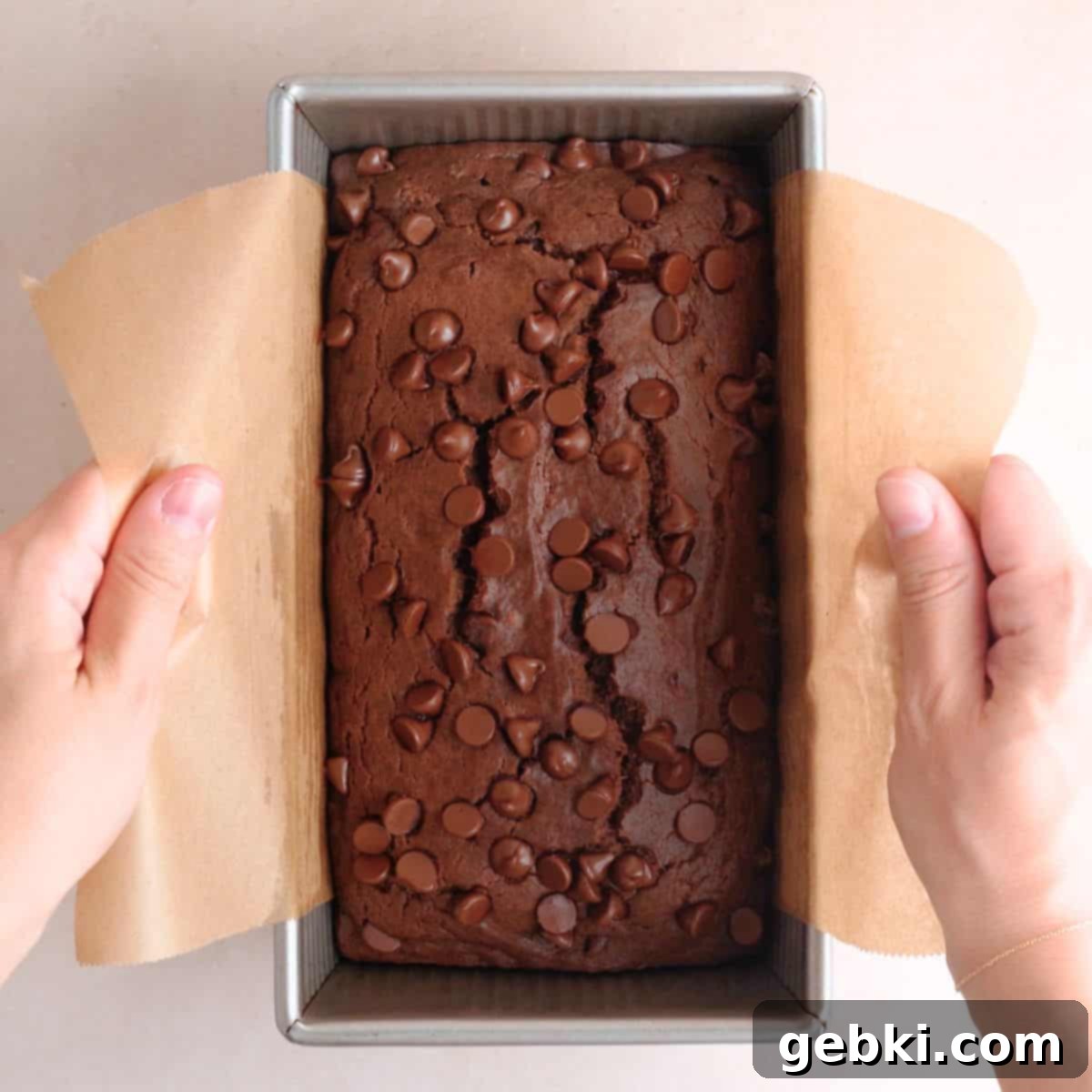 Decadent Double Chocolate Loaf 9 A freshly baked chocolate loaf cake being lifted out of its metal loaf pan by the parchment paper overhang, revealing its perfectly browned crust and moist interior.